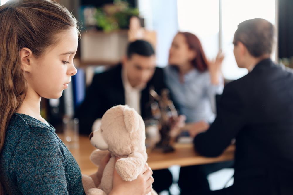 Sad young girl holding a teddy bear while adults argue in the background, depicting family conflict or divorce.