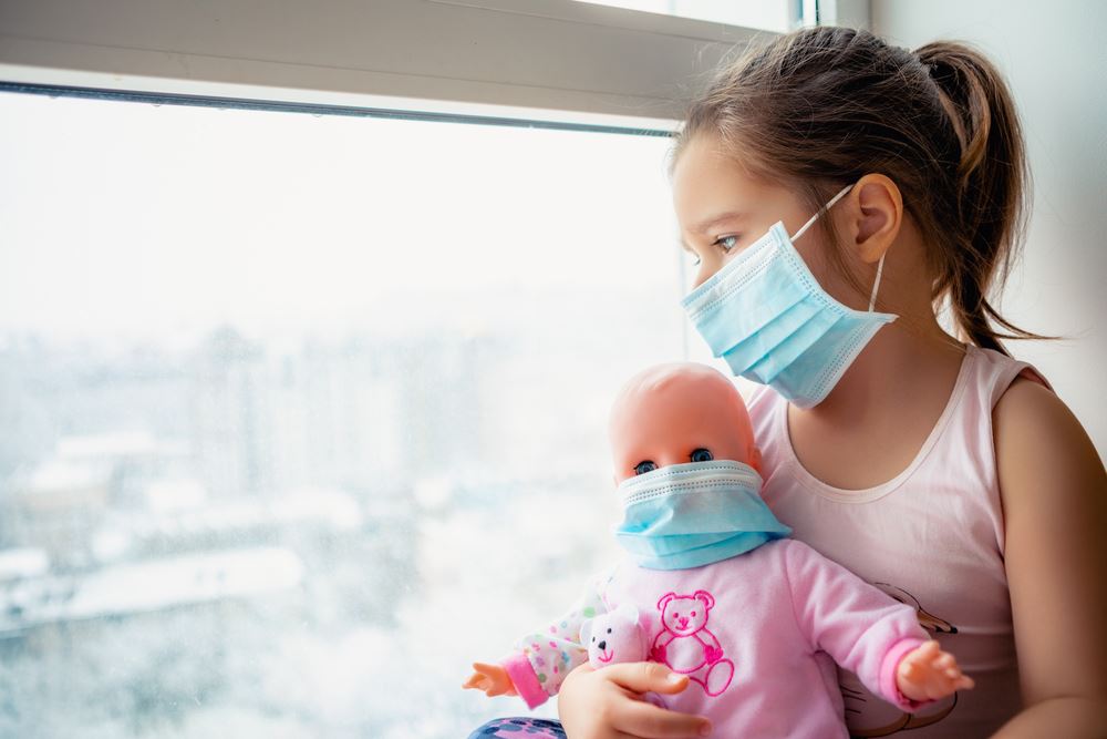 Young girl wearing a medical face mask holds a masked baby doll while gazing out a window.