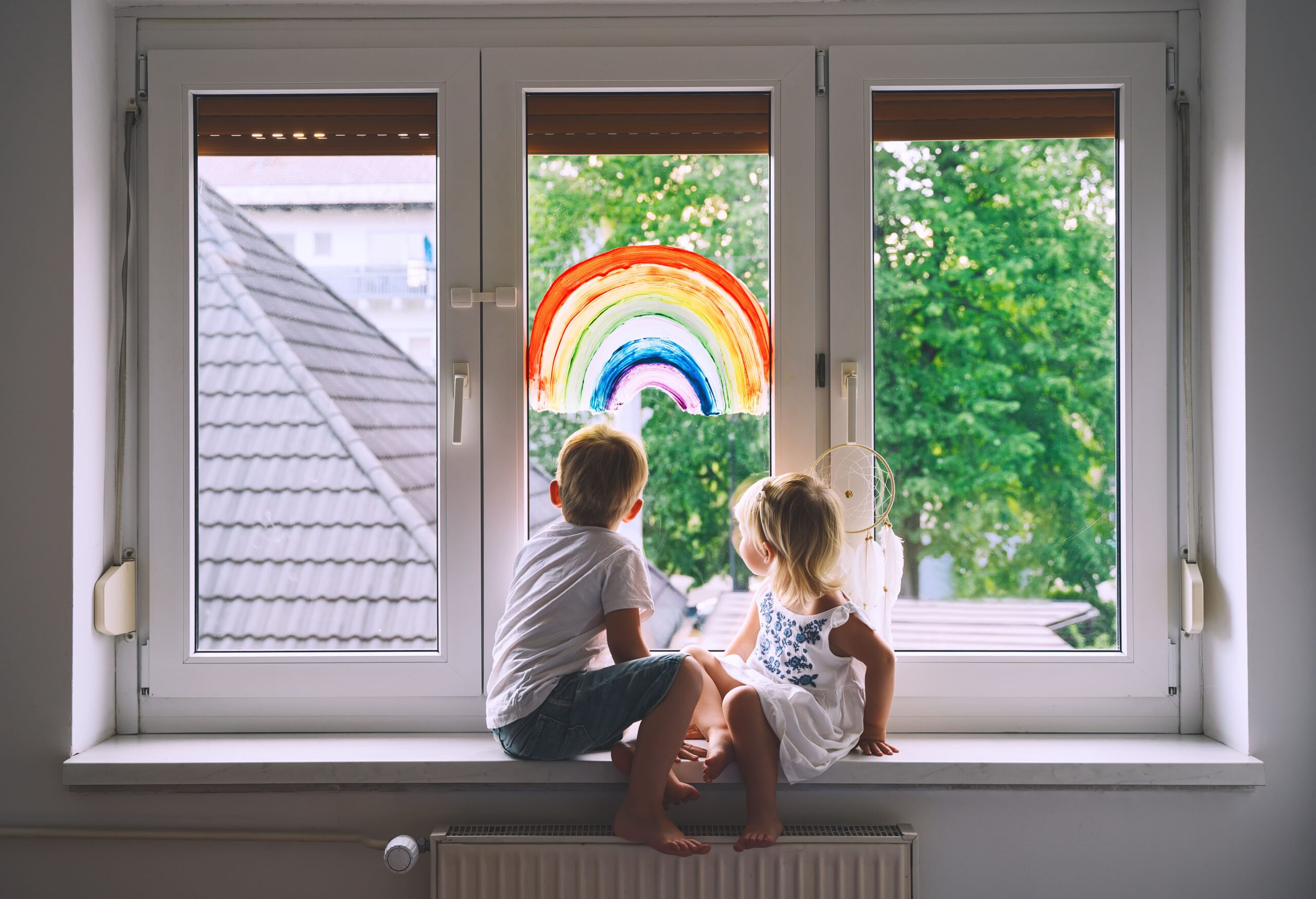 Two young children sitting on a windowsill looking outside at a colorful rainbow painting on the window glass.