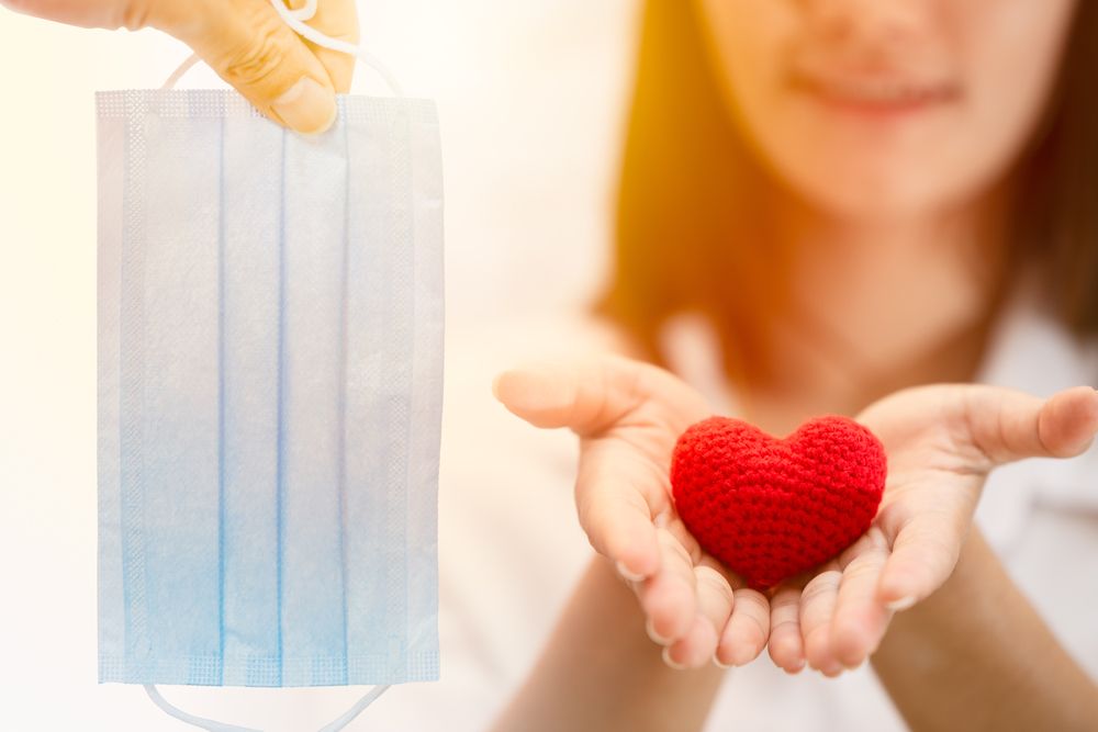 A smiling woman holds a red crocheted heart while a hand holds a blue surgical face mask nearby.