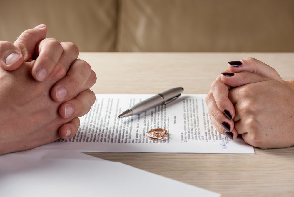 Two pairs of clasped hands on opposite sides of a divorce document with two wedding rings and a pen on top.