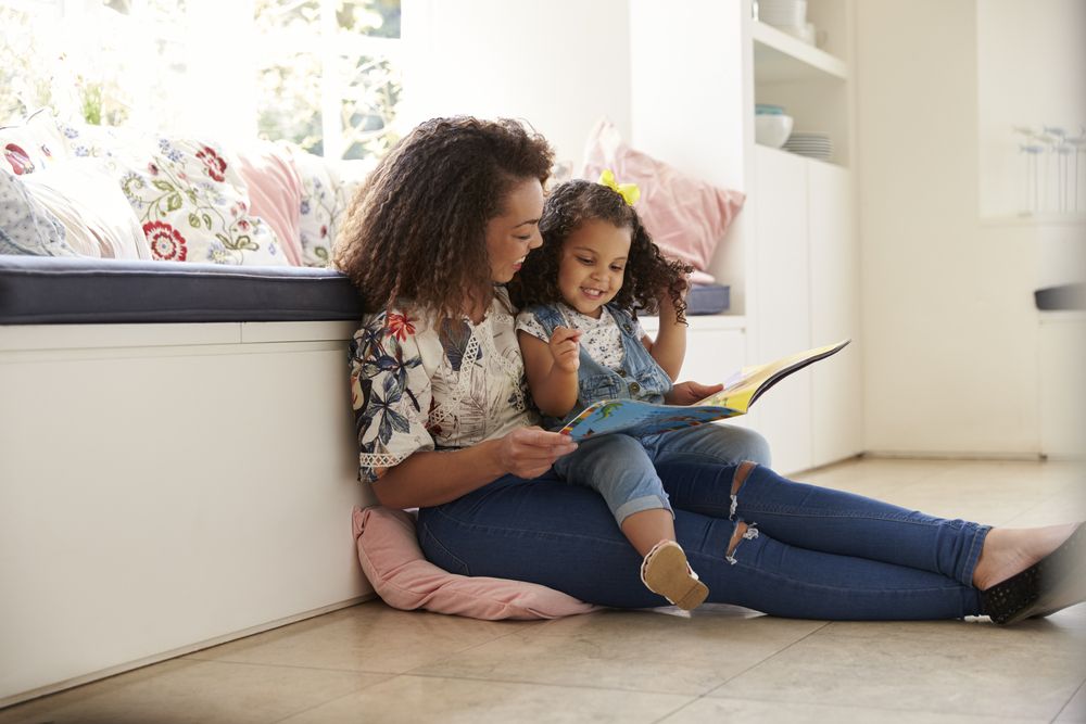 A mother and young daughter sitting on the floor together, reading a colorful picture book at home.