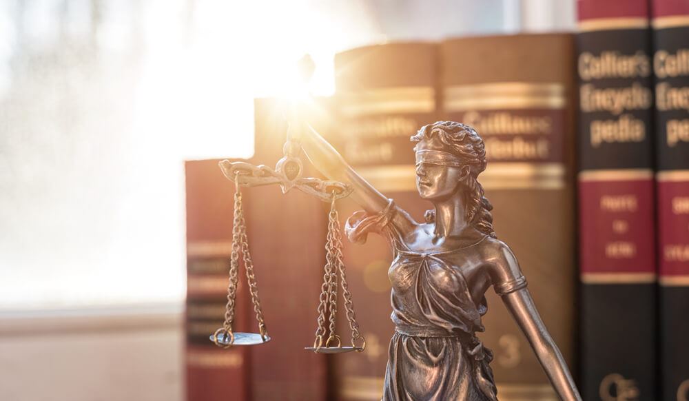 Bronze Lady Justice statue holding scales, blindfolded, with law books in the background and warm sunlight.