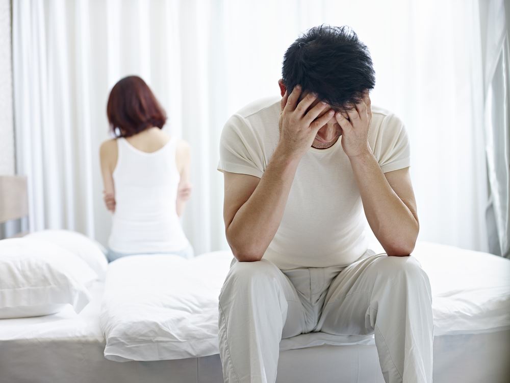 A distressed man sits on a bed with his head in his hands while a woman sits with her back turned in the background.