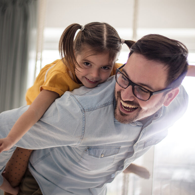 A laughing father with glasses gives his smiling young daughter a piggyback ride indoors.