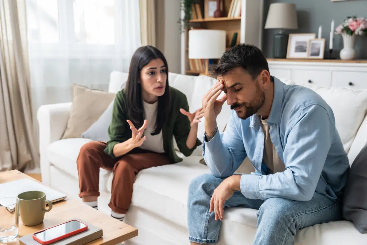 A woman gestures while arguing with a stressed man holding his head on a white couch in a living room.